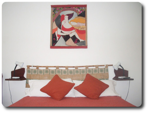 La foto muestra la cama matrimonial de una habitación doble de la hostería. En la pared cuelga un cuadro de un artista del lugar. Todo el lugar está decorado con trabajos de artistas y artesanos de San Marcos Sierras. Las habitaciones pueden ser dobles, triples, cuádruples o séxtuples.