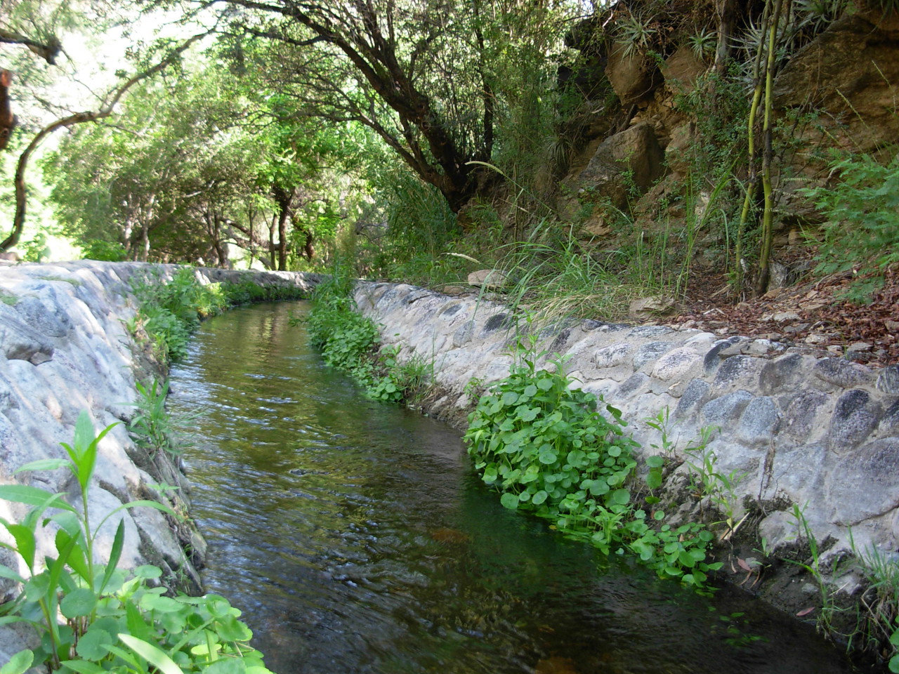 La foto muestra en primer plano una acequia cerca de la hosteria, uno de los lugares para disfrutar de las vacaciones, el descanso y la tranquilidad en el parque de la Hostería frente a las sierras del Cuniputo. La hostería le ofrece un buen alojamiento en San Marcos Sierras y un lugar con todas las comodidades para su relax.