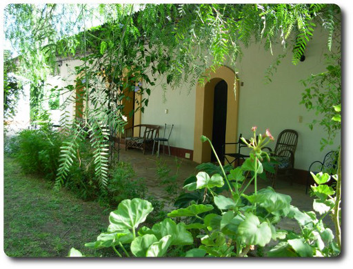 La foto muestra en el primer plano el ambiente tranquilo y natural lleno de vegetación del hospedaje, en un segundo plano se ven puertas de las habitaciones que dan a una galería.